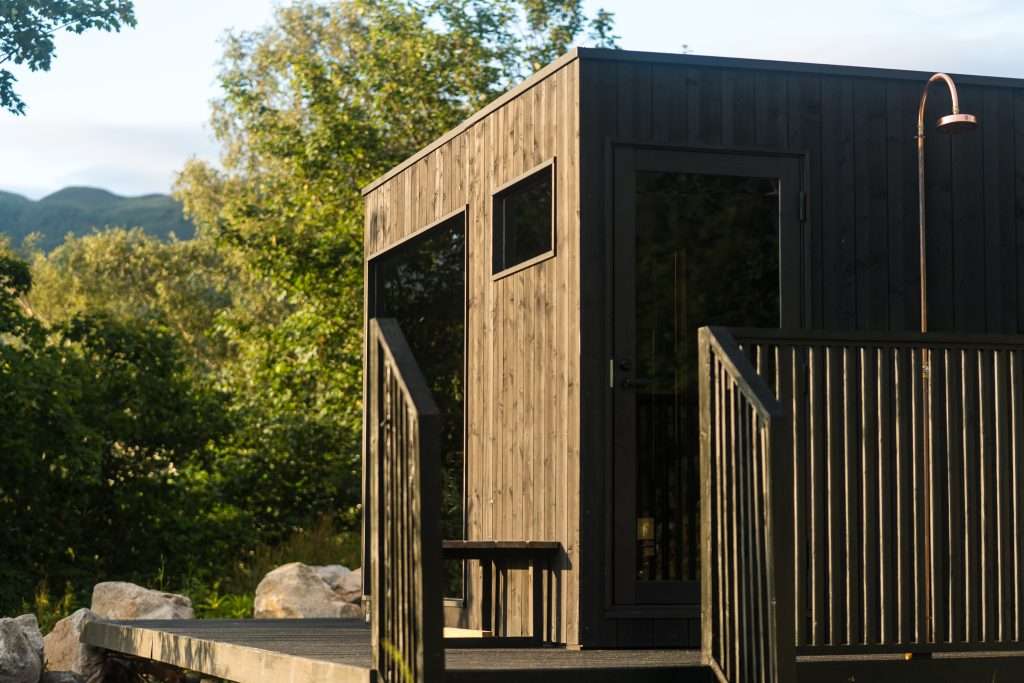 Woodlands Wilderness Sauna in Glencoe in focus. The building is a black cube made of vertical planks of wood.