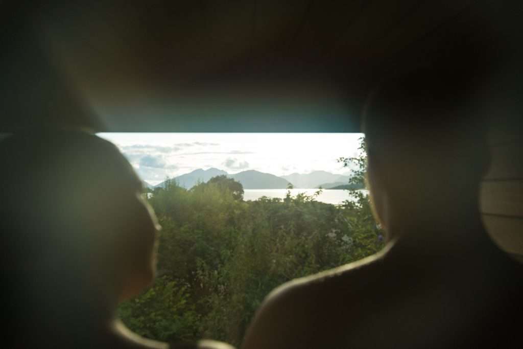 A couple inside the Woodlands Wilderness Sauna look out of the glass window at the panoramic view of the loch and mountains.