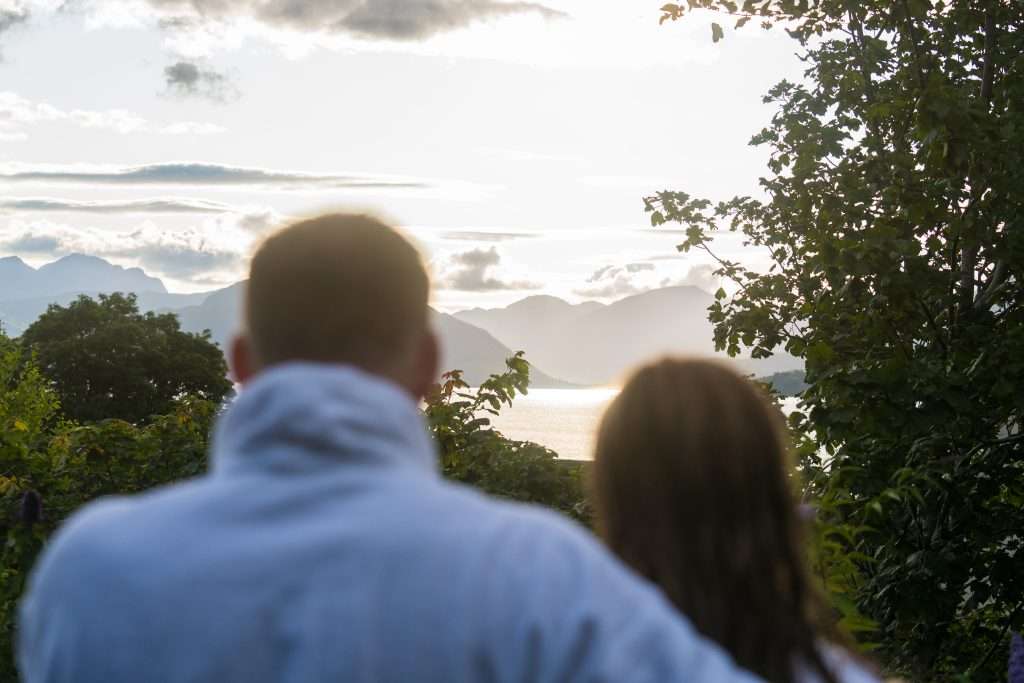 A couple outside the Woodlands Wilderness Sauna look out of the glass window at the panoramic view of the loch and mountains.