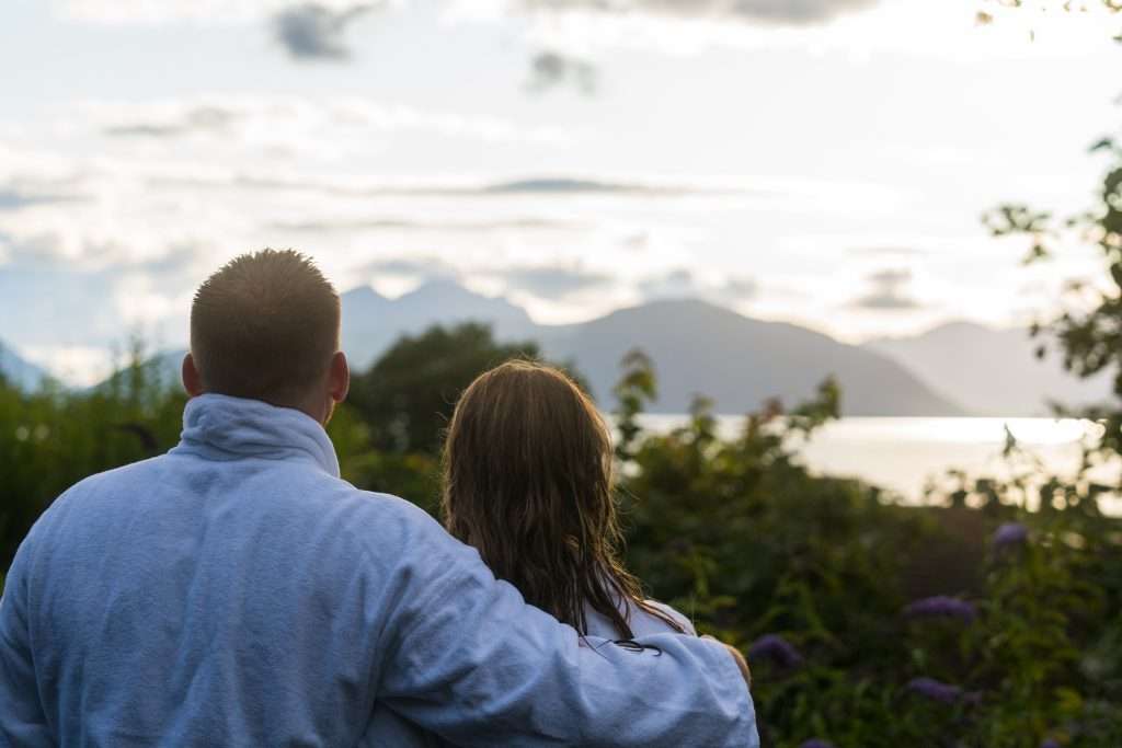 Massage, Wellness & Spa in Glencoe 10 A couple outside the Woodlands Wilderness Sauna look out of the glass window at the panoramic view of the loch and mountains.