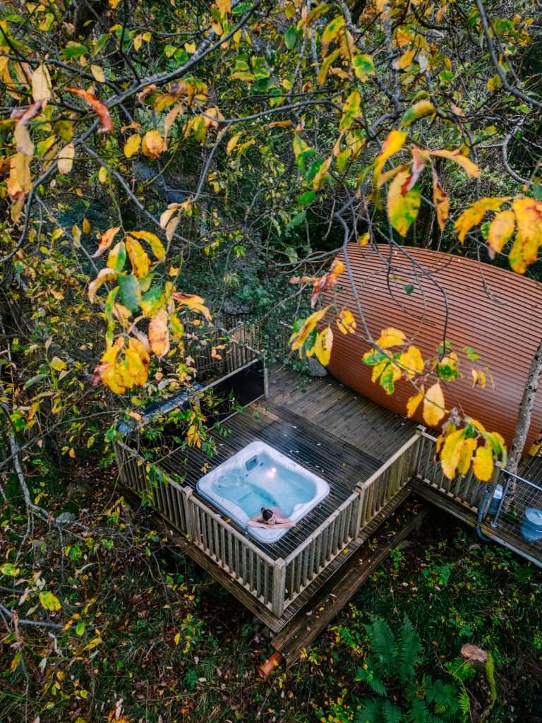 RiverBeds Luxury Wee Lodges from above. A lady lounges in the hot tub beneath the autumnal trees.