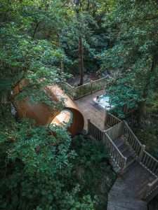RiverBeds Luxury Wee Lodges with Hot Tubs at Woodlands Glencoe. A view from through the forest towards the river with cabin and woman enjoying the bubbles.