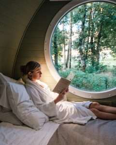 Relaxing at RiverBeds Luxury Wee Lodges with Hot Tubs at Woodlands Glencoe. A woman cosy in bathrobe reading a book on quality bedlinen and forest view.
