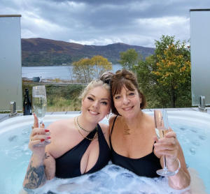 Mother and daughter enjoy champagne in the hot tub at SeaBeds Luxury Lookout Lodges at Woodlands Glencoe with mountain and loch view. Perfect mothers day destination.