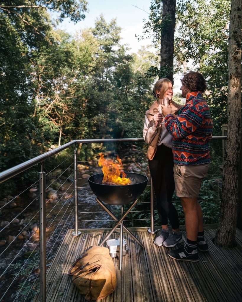 A couple clink champagne glasses by the roaring fire pit on the riverside at the romantic RiverBeds Luxury Wee Lodges with Hot Tubs at Woodlands Glencoe.