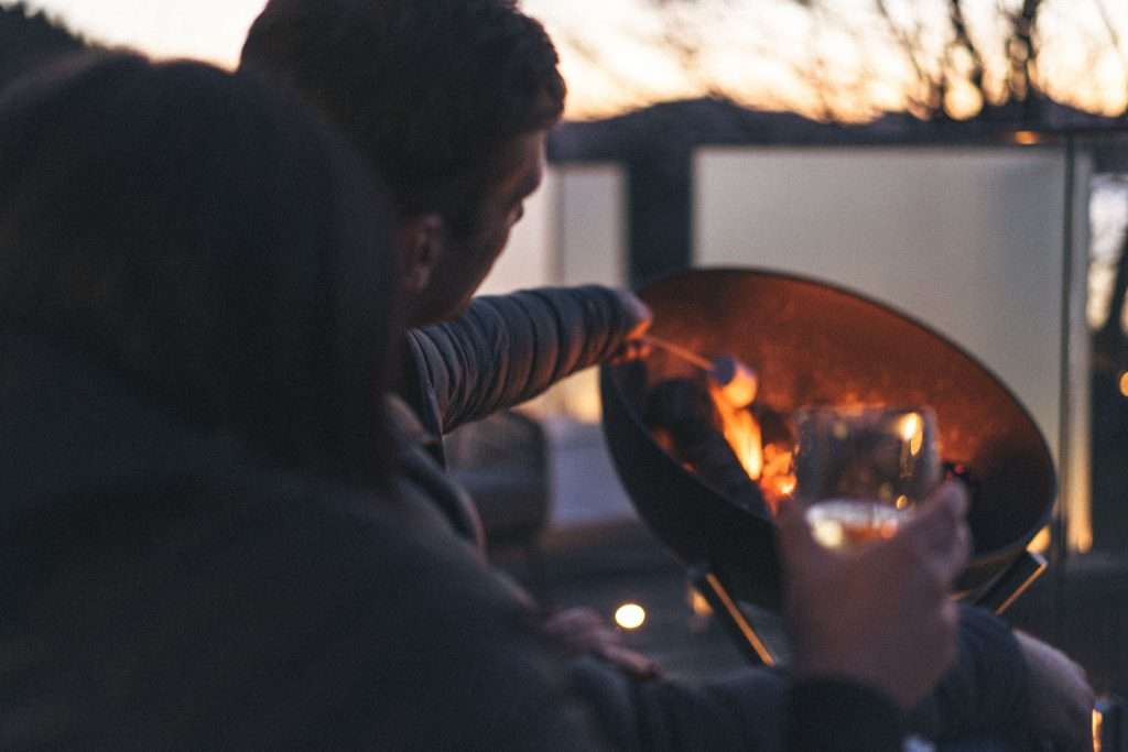 A romantic couple sip wine and toast marshmallows on the fire pit at sunset at SeaBeds Luxury Lookout Lodges with Hot Tubs at Woodlands Glencoe.