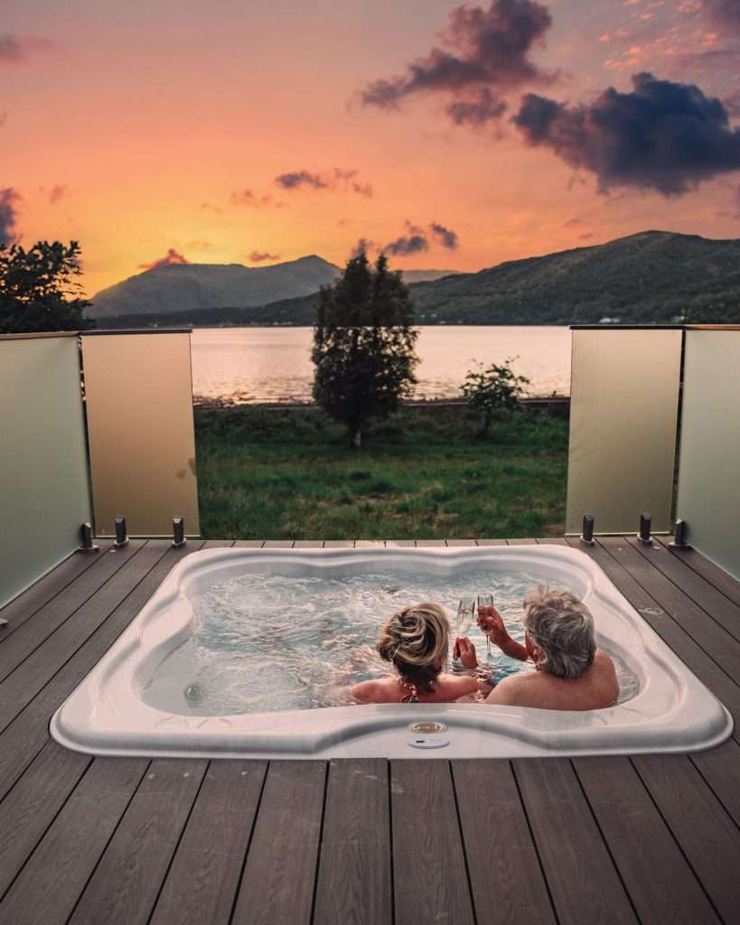 A couple clink champagne glasses and enjoy the sunset in the hot tub on the private terrace of SeaBeds Luxury Lookout Lodge at Woodlands Glencoe.