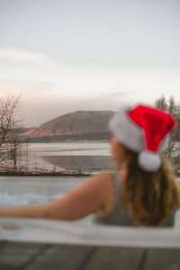 Romantic christmas getaways at SeaBeds Luxury Lookout Lodges with Hot Tubs at Woodlands Glencoe. A woman sits in the hot tub wearing a santa hat looking out over the loch and mountain view.